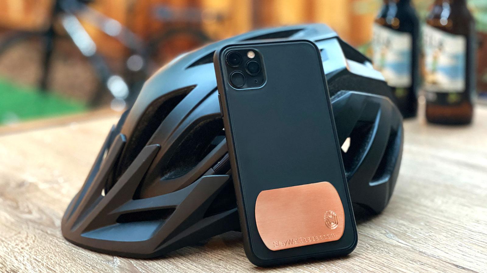 Phone case with 2" x 1.5" Adhesive Phone Patch made in USA from natural antimicrobial copper attaches to germy devices we touch most. Phone case with phone patch shown leaning up against a cyclist helmet. Sanitizers, Chemical Free, Lasts Forever. Recyclable. Kills 99.9% of Harmful Bacteria. EPA registered.