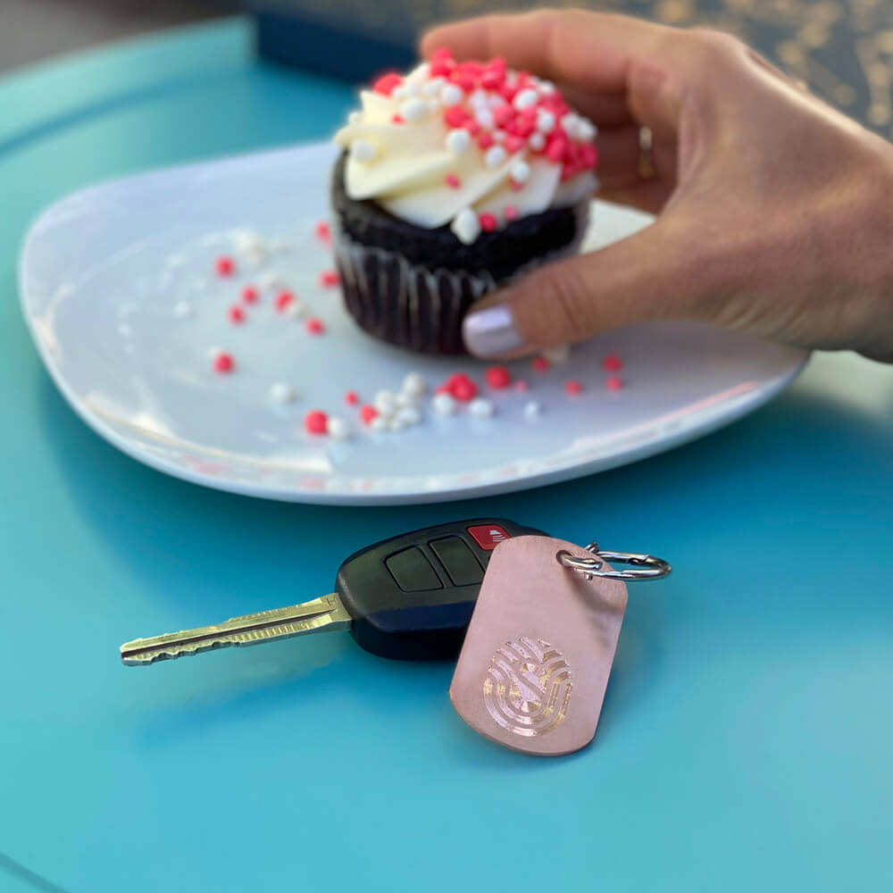 Lady's hand with polished nails grabbing a chocolate cupcake with white frosting and pink and white sprinkles. Next to plate is a car key on a chain along with a Copper dog tag with StayWell logo  kills 99.97% of germs and bacteria!