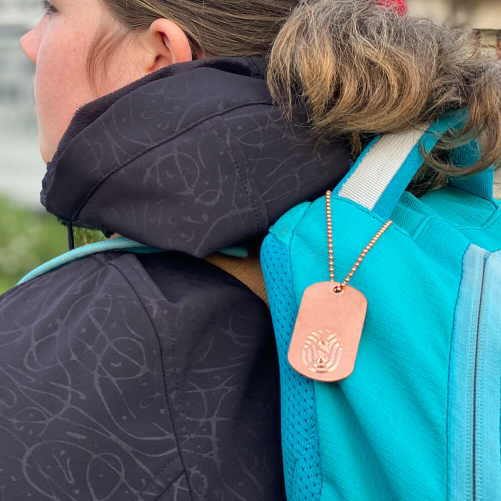 Staywell Copper dog tag hanging on bed chain on turquoise blue backpack on girl with black warm hooded jacket. She has a pony tail and brown hair.